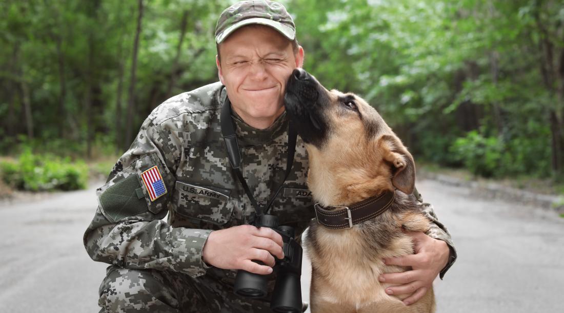 A service member in uniform kneeling down as a German Shepherd dog is giving him a kiss on the cheek.